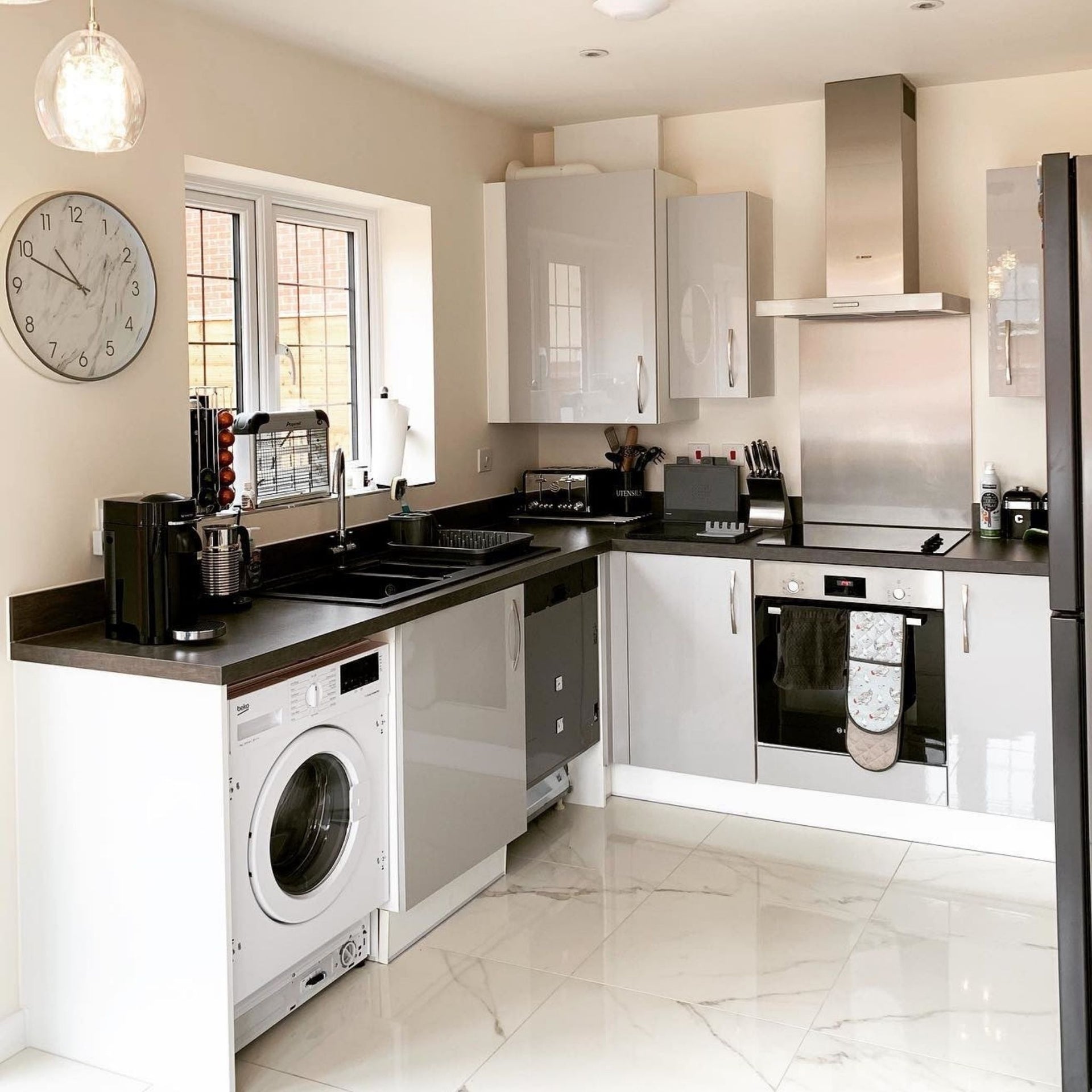 A modern kitchen interior with firenze statuario white marble effect floor tiles, white and silver cabinets, dark countertops, a built-in washing machine, dishwasher, and oven, plus a large round wall clock and pendant light.
