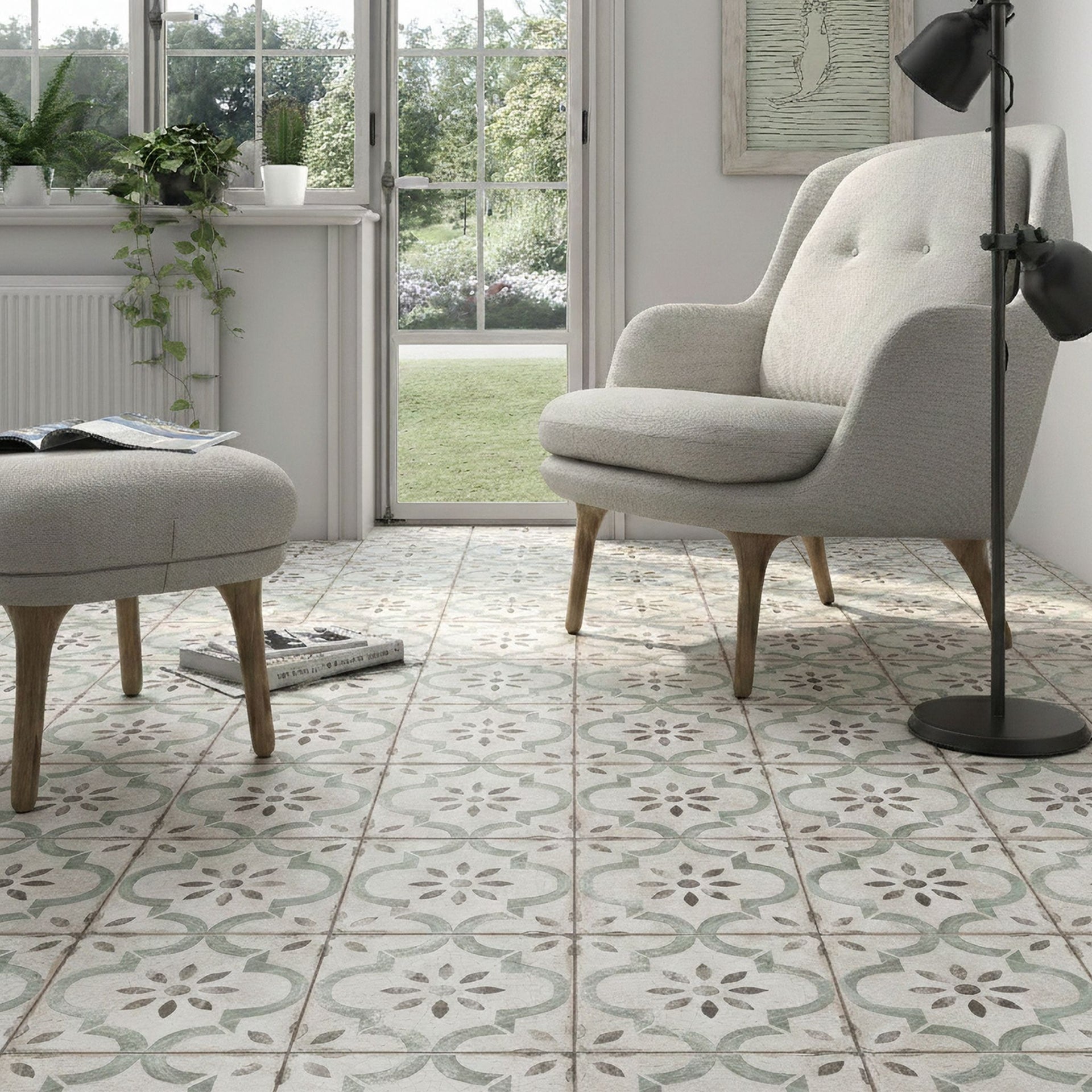 Bright conservatory living room featuring a grey armchair with a matching ottoman, positioned on classic green patterned floor tiles. Large windows offer a clear view of the outdoor greenery.