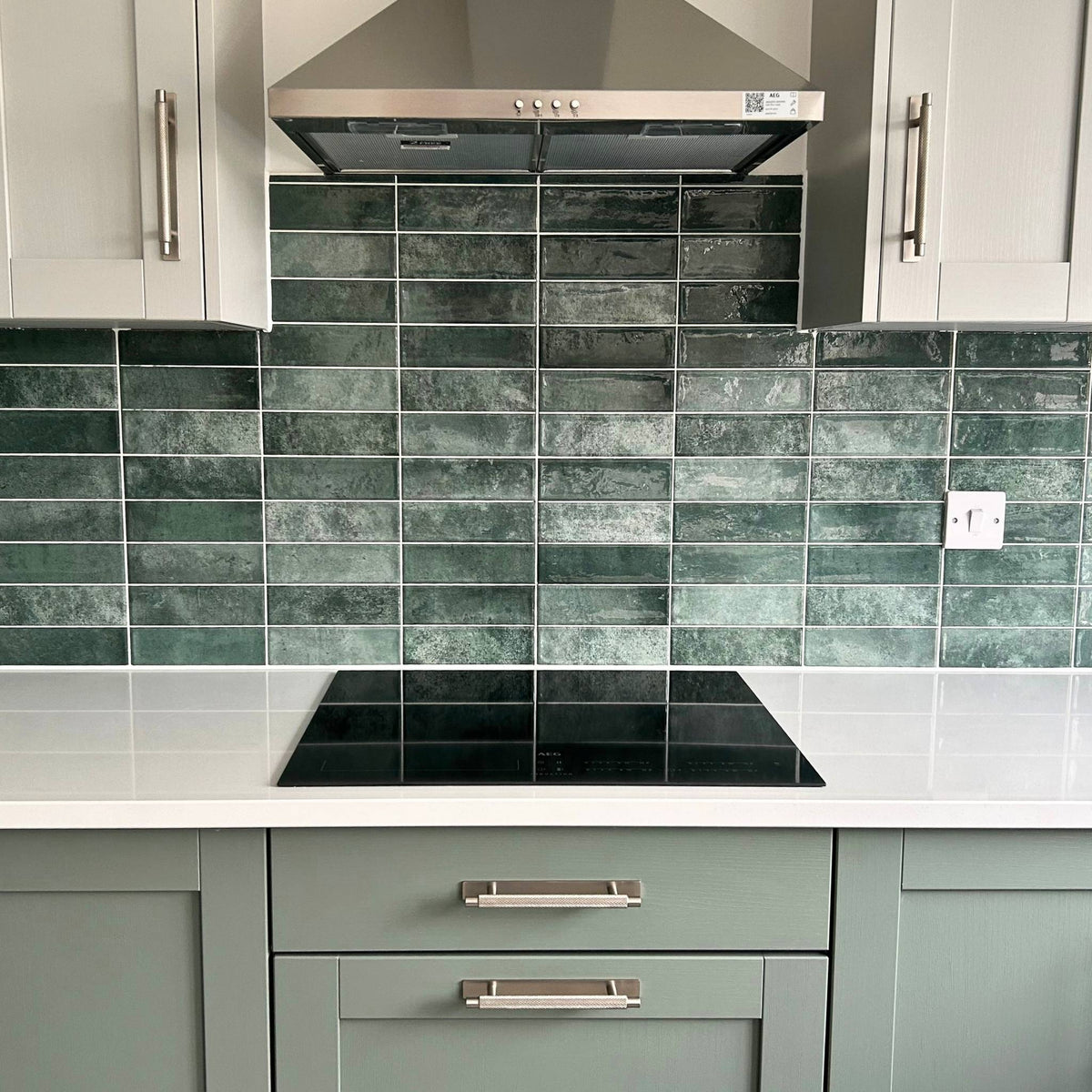 A modern kitchen backsplash featuring Loxwood glossy rectangular green tiles with a textured, stone-like finish, paired with sage green cabinets, a white countertop, and a black built-in cooktop.


layout-kitchen