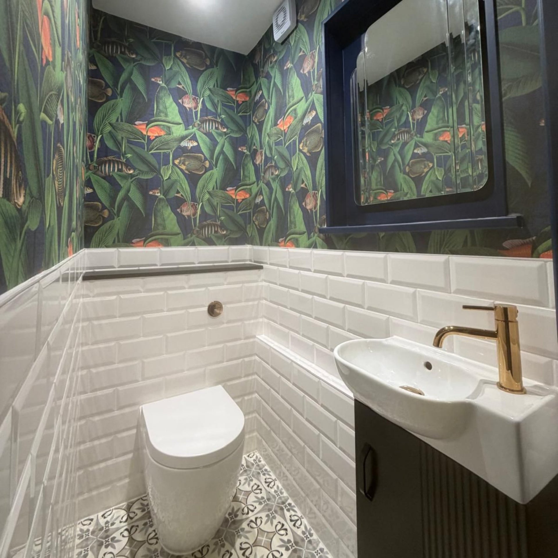 Bathroom with botanical wallpaper, white tiled brick walls, a toilet, and a sink with gold faucet.