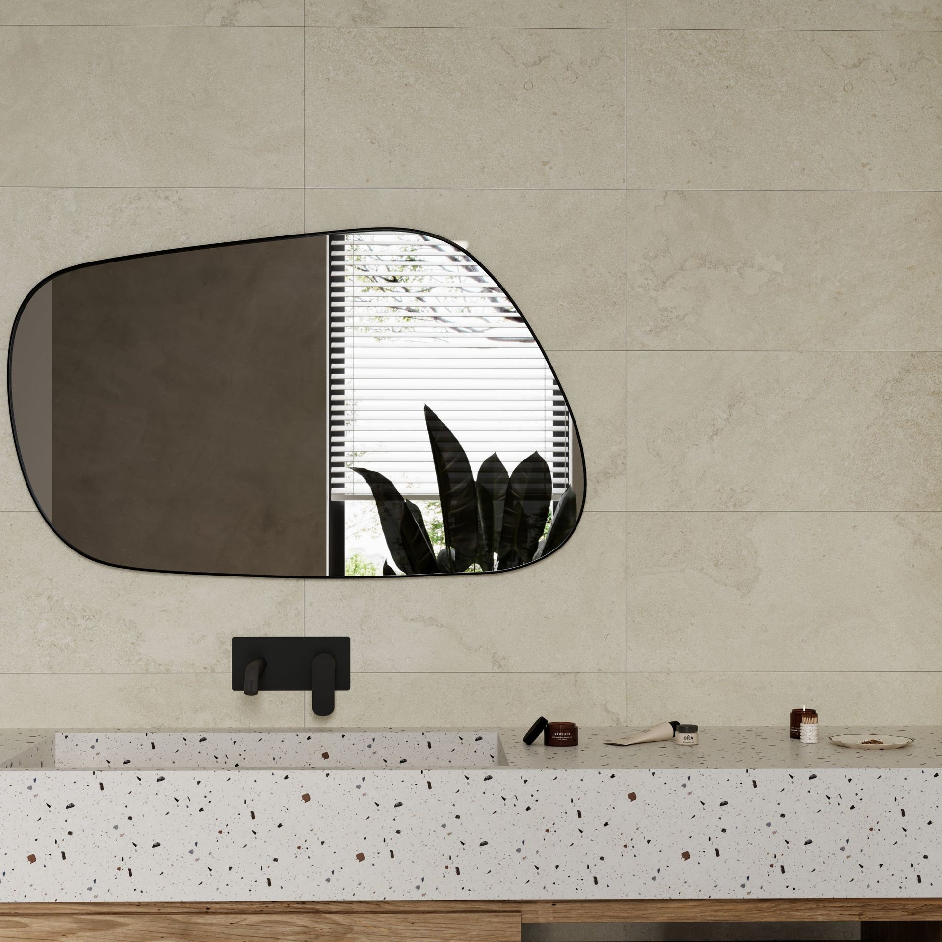 An organic-shaped wall mirror with a dark frame is mounted on an Oxton bone beige stone effect bathroom wall tile, above a white speckled vanity countertop with a black faucet.