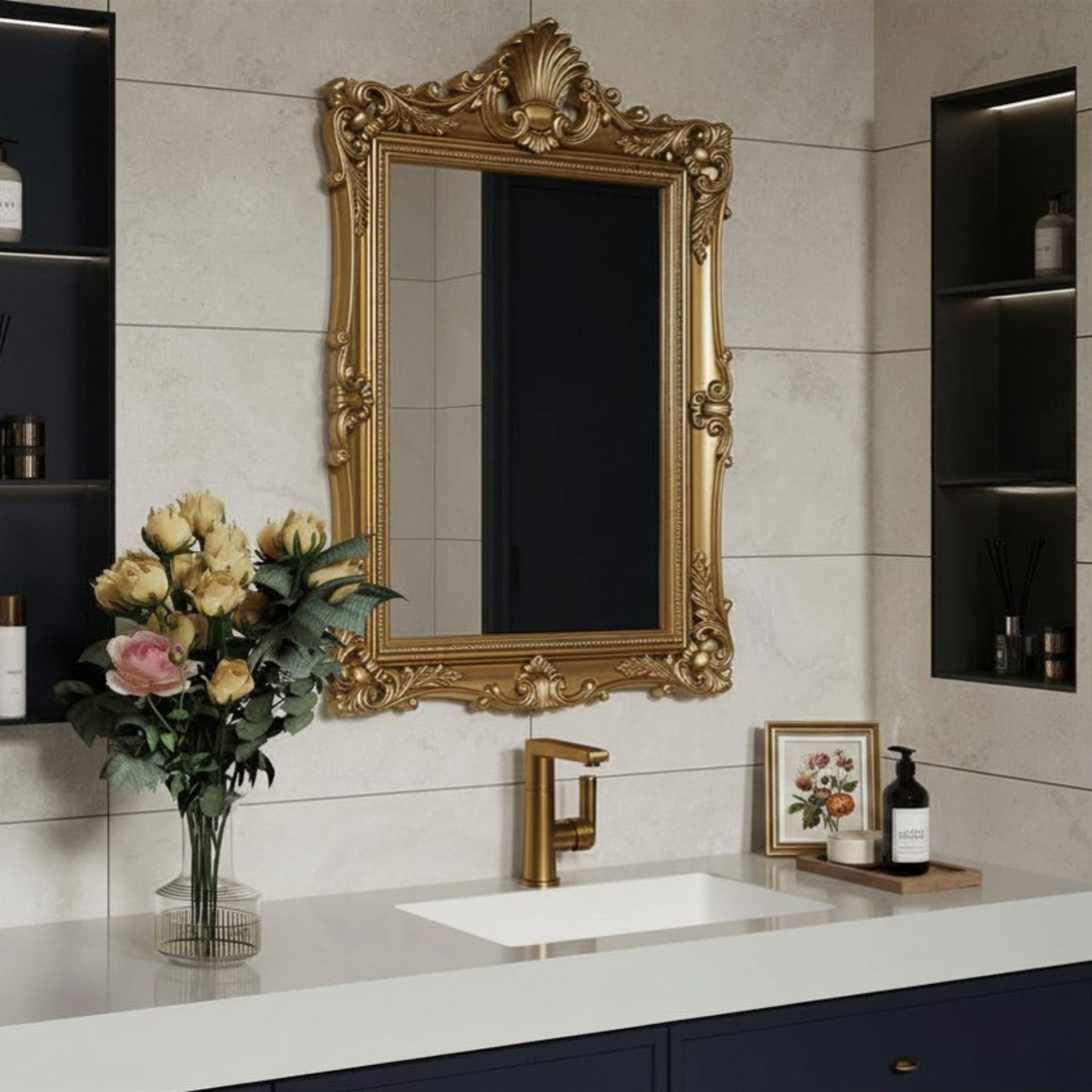An ornate gold-framed rectangular wall mirror hangs above a white bathroom vanity with a gold faucet, set against off white Oxton stone effect tile walls with dark shelving on either side.
