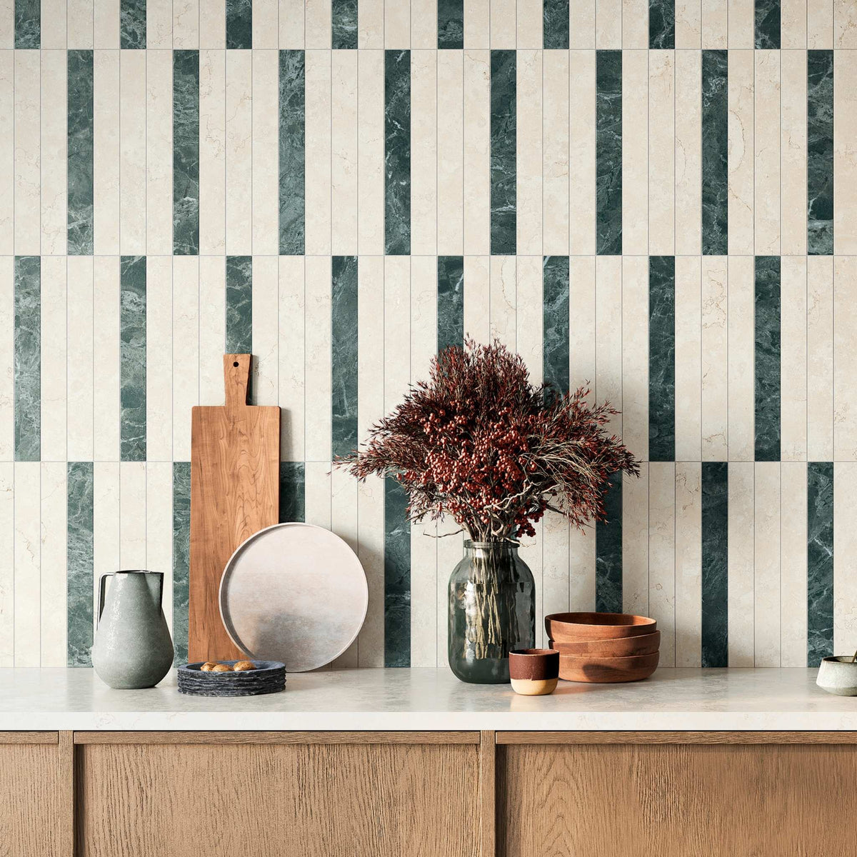 A styled kitchen countertop featuring a vertical striped backsplash , using alternating Sillara White and Green marble and stone-look tiles. The counter is decorated with a gray ceramic pitcher, a wooden cutting board, a round plate, a dark glass vase filled with dried burgundy foliage, and stacked wooden bowls.

