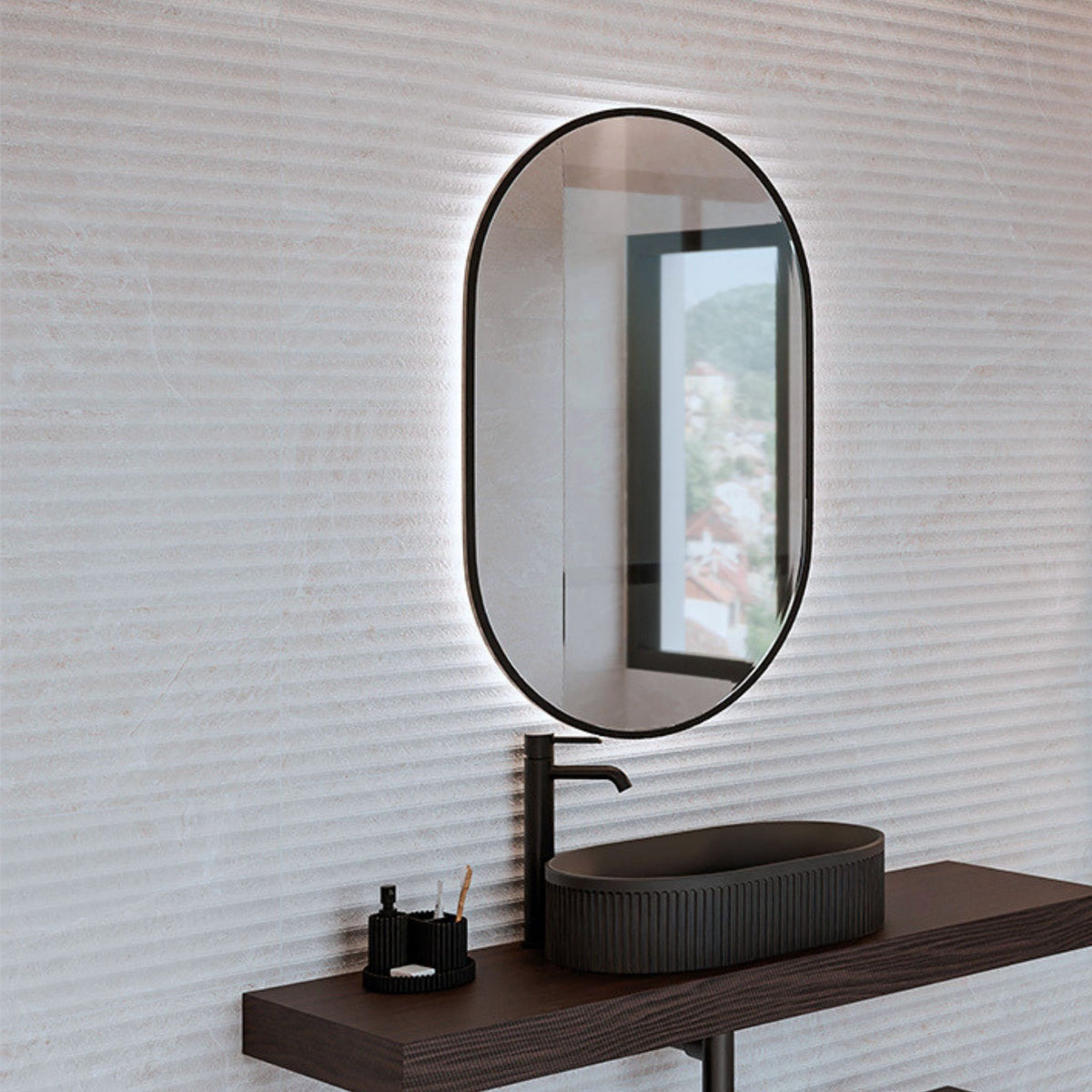 An oval LED-backlit bathroom mirror with a black frame, mounted on a Zamora White fluted textured decorative stone-look wall tiles above a dark wood vanity with a dark vessel sink.